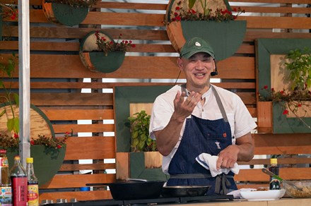 An Asian man in white shirt and denim apron, wearing a headset microphone, smiles and gestures while standing over a stove.