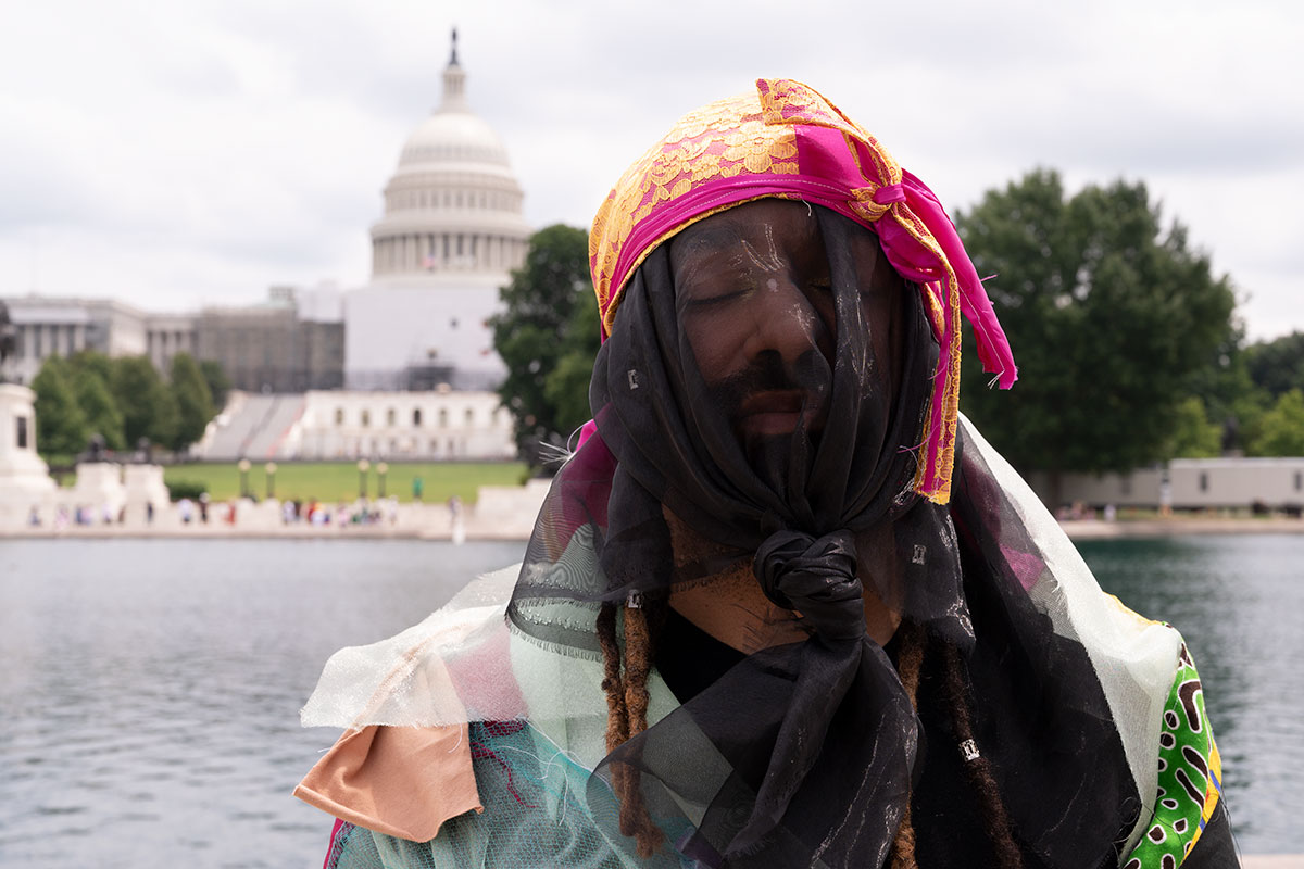 A Black person wears a bright pink and yellow durag with a black veil tied under their chin and quilted fabric scraps draping over their shoulders. Their eyes are closed. In the background, the U.S. Capitol Building.