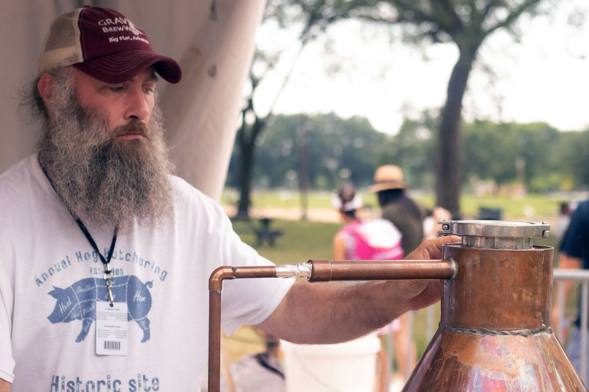 A man with a big gray beard and Annual Hog Butchering T-shirt inspects a copper still outdoors.