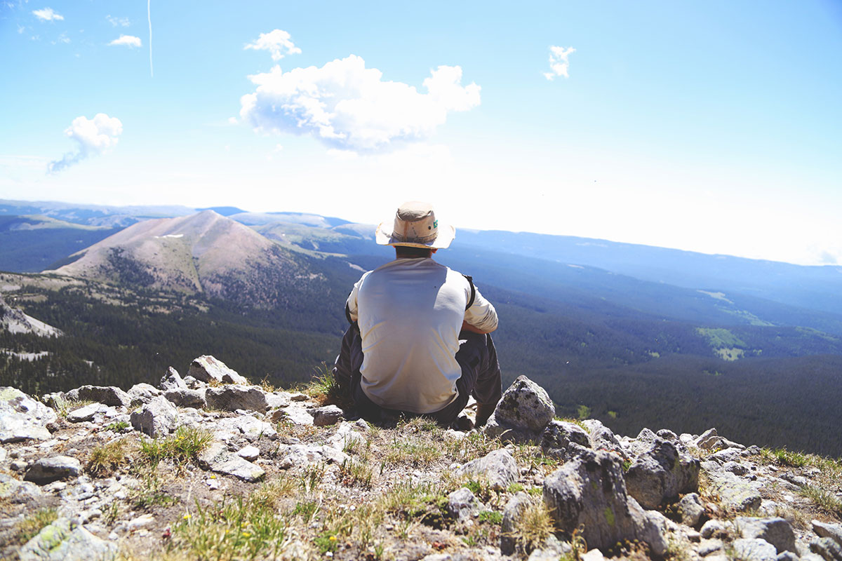 From behind, a person sits on the edge of a mountain or ridge top, looking out at expansive landscape, including rolling hills and white clouds in a blue sky.